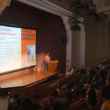 a person speaking on stage in front of a podium with people seated in the audience