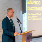 A person in a blue suit stands at a lectern to give a presentation in front of a projection screen