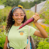 A child in a green shirt and pink glasses holds up a large red carrot in a garden