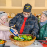 Three people work together to mix a salad in a large wooden bowl