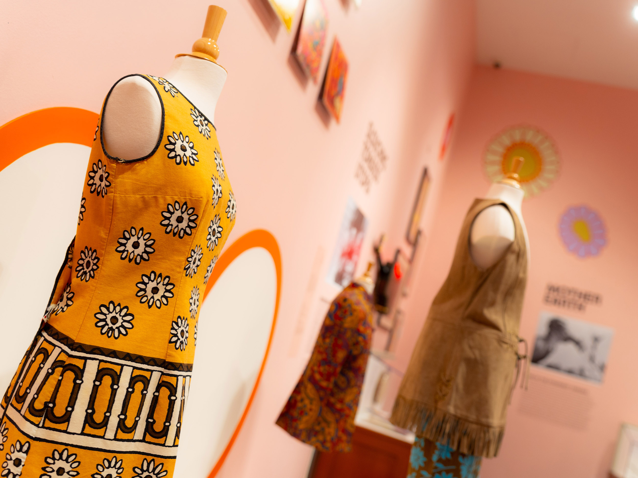 An arrangement of wall art and dress forms displaying yellow, red, and brown outfits from the 1960s