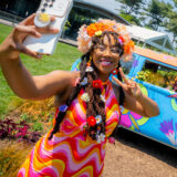 A person in a colorful yellow and pink outfit takes a selfie in front of a blue VW bus