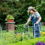 A person wearing a baseball cap and overalls uses a walker to navigate a green garden
