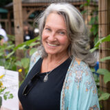 A person with long silver hair, wearing a blue shawl, black shirt, and sunglasses hung from their collar pose for a photo