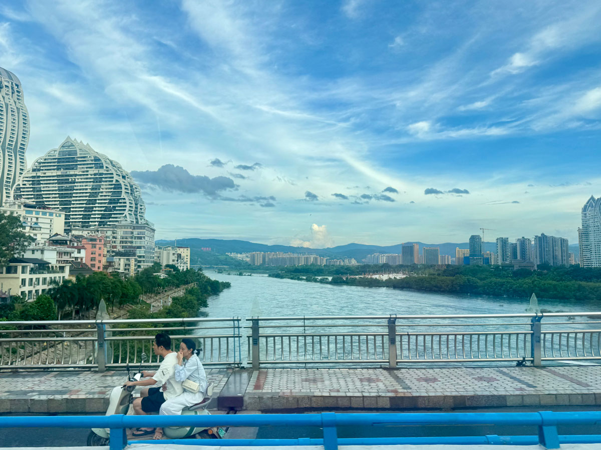 Two people ride a moped in front of a river with a city skyline in the background.