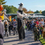 A tall skeleton puppet parades along a path lined with children and adults in festive clothing