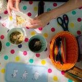A person's hands can be seen mixing up a packet of tea leaves among a polka-dot covered table featuring pots of plant material and an orange basket of colored pencils