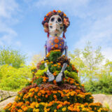 A tall, stylized sculpture of a skeleton, wearing a dress of orange and purple flowers and a flower crown, under a sunny blue sky