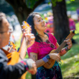 Two people perform with small acoustic guitars outdoors on a sunny day