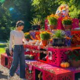 A person in blue jeans and a long-sleeved white shirt places a memento on a tall altar of pink and orange cubes, covered in notes, photos, and potted green plants