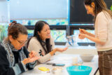 two people sitting at a table with two bowl in front of them and another person standing up holding a utensil
