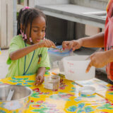 A child in a green shirt uses a spoon to mix spices in a plastic bag