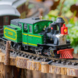 A green toy locomotive moves along a track lined with green vegetation