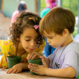 Two children work together to pot up seedlings in green plant pots