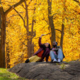 Two people in warm fall clothes sit on a rocky outcrop, taking a selfie together among bright, golden-hued trees and green grass