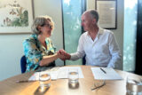 Two people shake hands in a sunny conference room setting, with papers and pens laid out before them