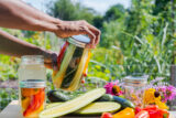 two hands holding a glass jar containing sliced vegetables in liquid in front of cucumbers, peppers, and flowers on a wood surface
