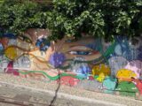 A colorful mural on a cement wall beneath an overhanging fringe of green foliage