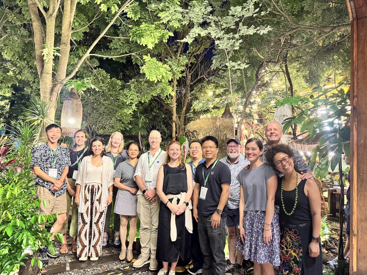 A group of people stand together in front of leafy green trees.