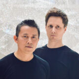 Two people with short hair, wearing black shirts, pose for a photo in front of a shimmering white wall