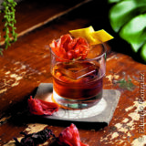 A colorful cocktail in a glass tumbler sits among green plants