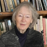 A person with gray hair, wearing a black sweater, poses for a headshot in a library setting