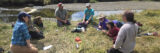 A group of people sit in the grass while discussing something outdoors on a sunny day, with forest visible in the background