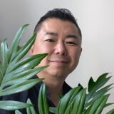 A person with short black hair peeks out from behind a number of green tropical plant leaves