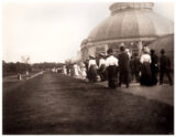 A group of people in white shirts and black skirts, and others in black suits, stroll alongside a conservatory dome in this black and white antique photo