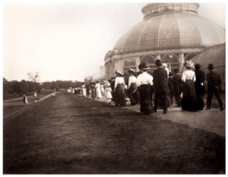 A group of people in white shirts and black skirts, and others in black suits, stroll alongside a conservatory dome in this black and white antique photo