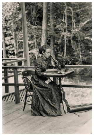 A person in a long dress sits on a wooden deck, looking into a microscope, with a forest visible in the background