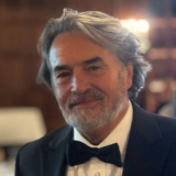 A person with middle-length gray hair, a salt-and-pepper-beard, and wearing a black and white tuxedo poses for a photo indoors