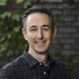 A person with short hair, wearing a black shirt, poses for a photo against a brick wall laced with climbing green vines