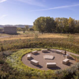 A person sits among a ring of flat stones, arrayed like benches around another central, rectangular stone. Surrounding the space are meadows of wild grasses and other plants, with an arid landscape visible in the background
