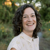 A person in a cream-colored shirt, with long, wavy dark hair, poses for a photo in a green outdoor space at sunset