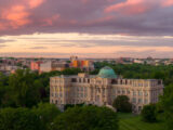 A classical building with a green dome sits under a sunset sky, surrounded by forest