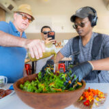 Three people mix up a large bowl of salad together in a kitchen