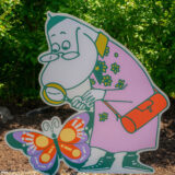A Moomin character in a pink outfit uses a magnifying glass to examine a colorful butterfly