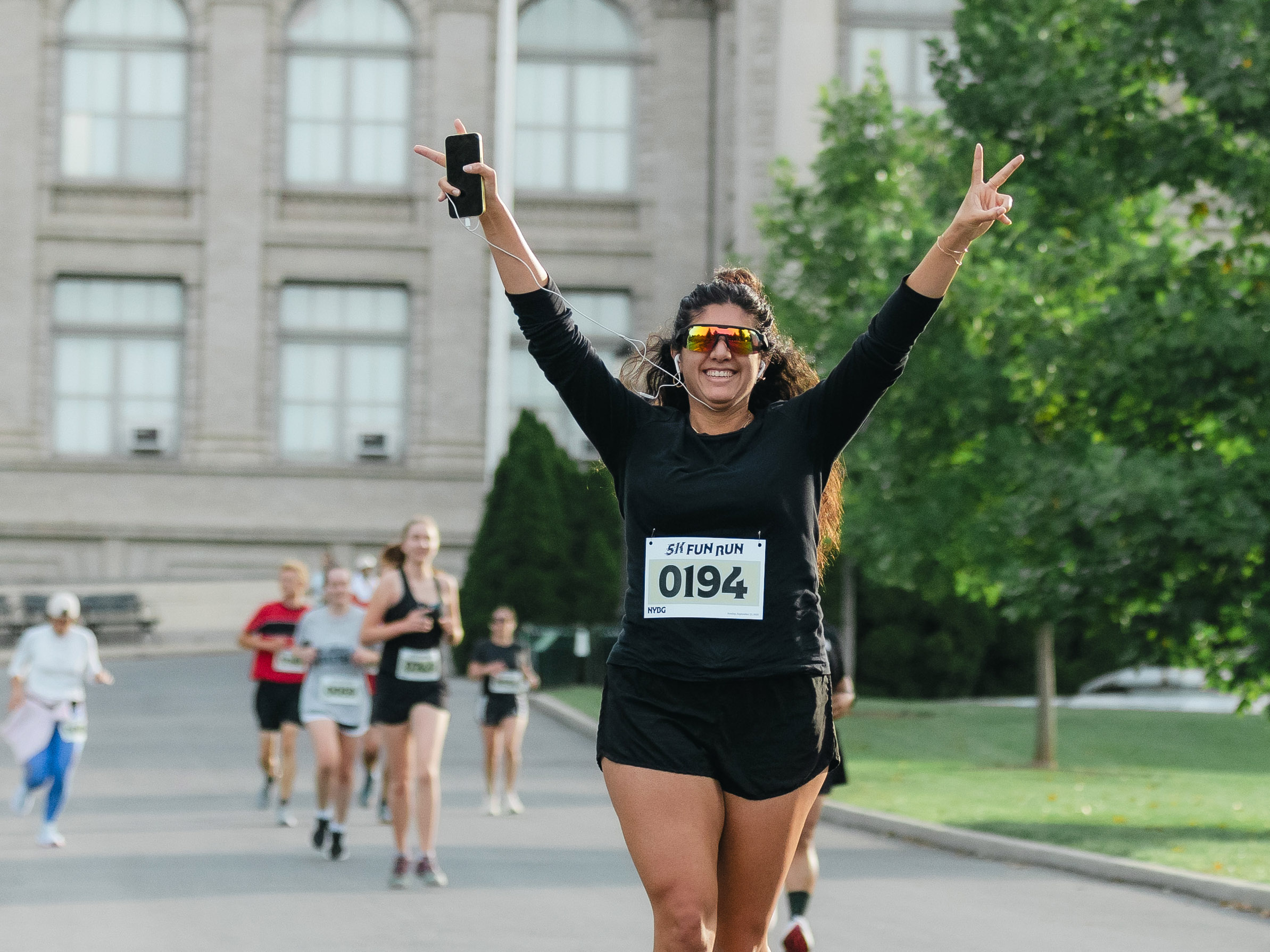 A person in black clothing throws peace signs in the air while running with a group on a paved path