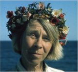 A film photo of a person with short blonde hair wearing a colorful flower crown, with the ocean visible beneath a blue sky in the background
