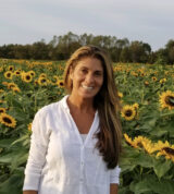 A person with long blonde hair, wearing a white shirt, stands in a field of yellow flowers
