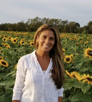 A person with long blonde hair, wearing a white shirt, stands in a field of yellow flowers