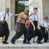 A group of dancers in black and white outfits dance in a line