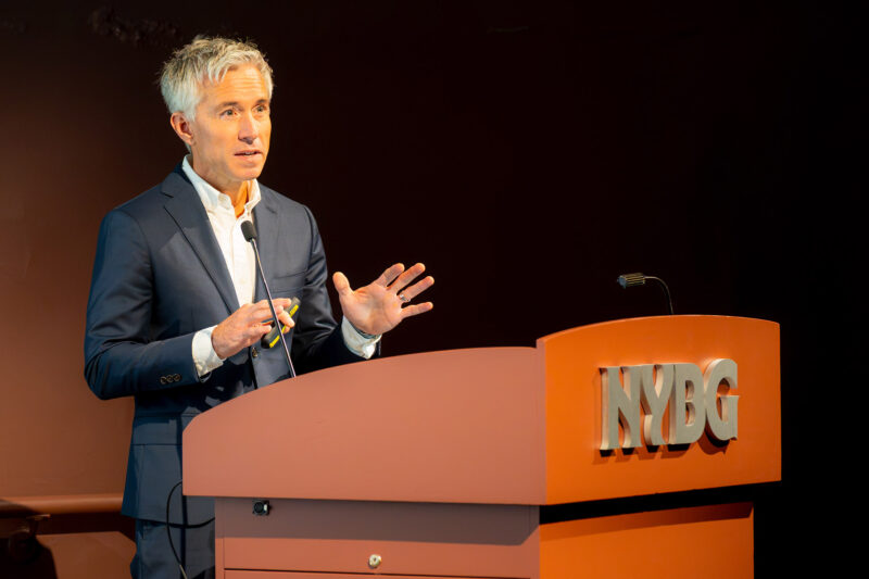 A person in a blue suit and white shirt speaks from a spotlit brown lectern