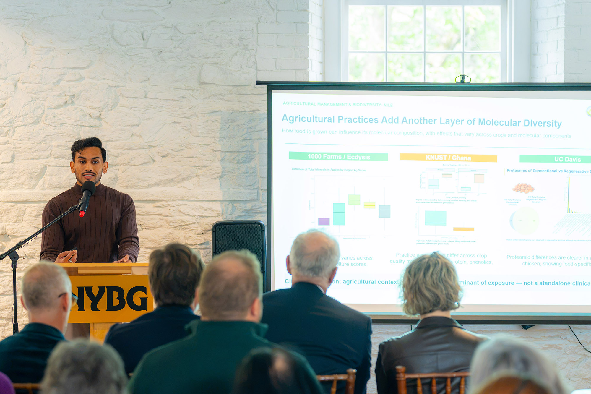 A person speaks at a lectern, presenting a projector screen presentation to a room of seated people