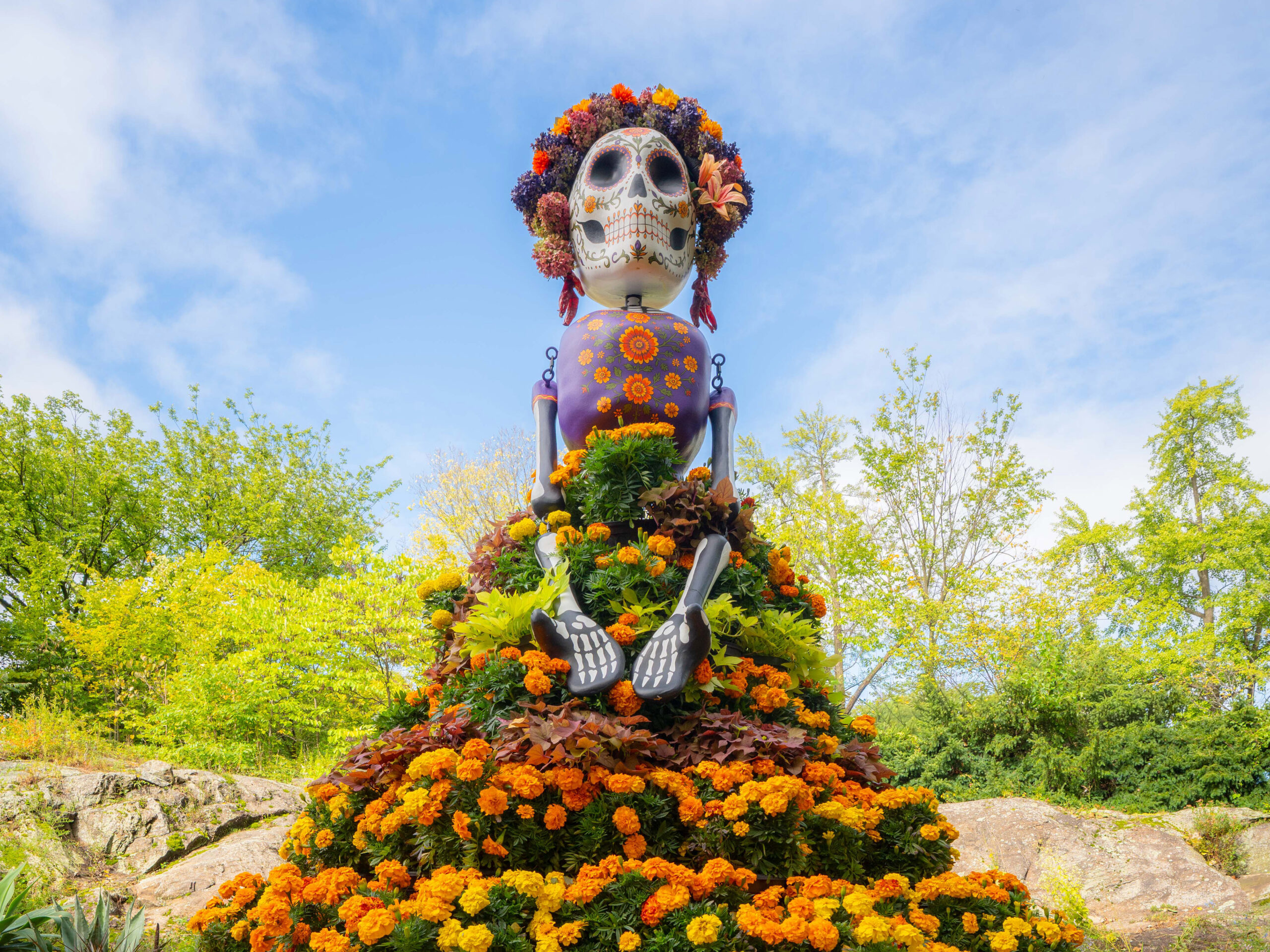 A tall, stylized sculpture of a skeleton, wearing a dress of orange and purple flowers and a flower crown, under a sunny blue sky