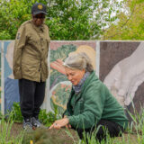 A person with long hair and a green sweatshirt kneels down to point at a plant, while a standing person, wearing a black baseball cap and brown coat, looks on