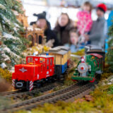 Red and green model trains travel along toy tracks as people look on in the background