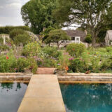 A cement foot bridge passes over a blue pool of still water toward a lush green garden on the other side