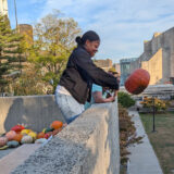 A person in blue jeans and a black jacket lobs an orange pumpkin off a roof